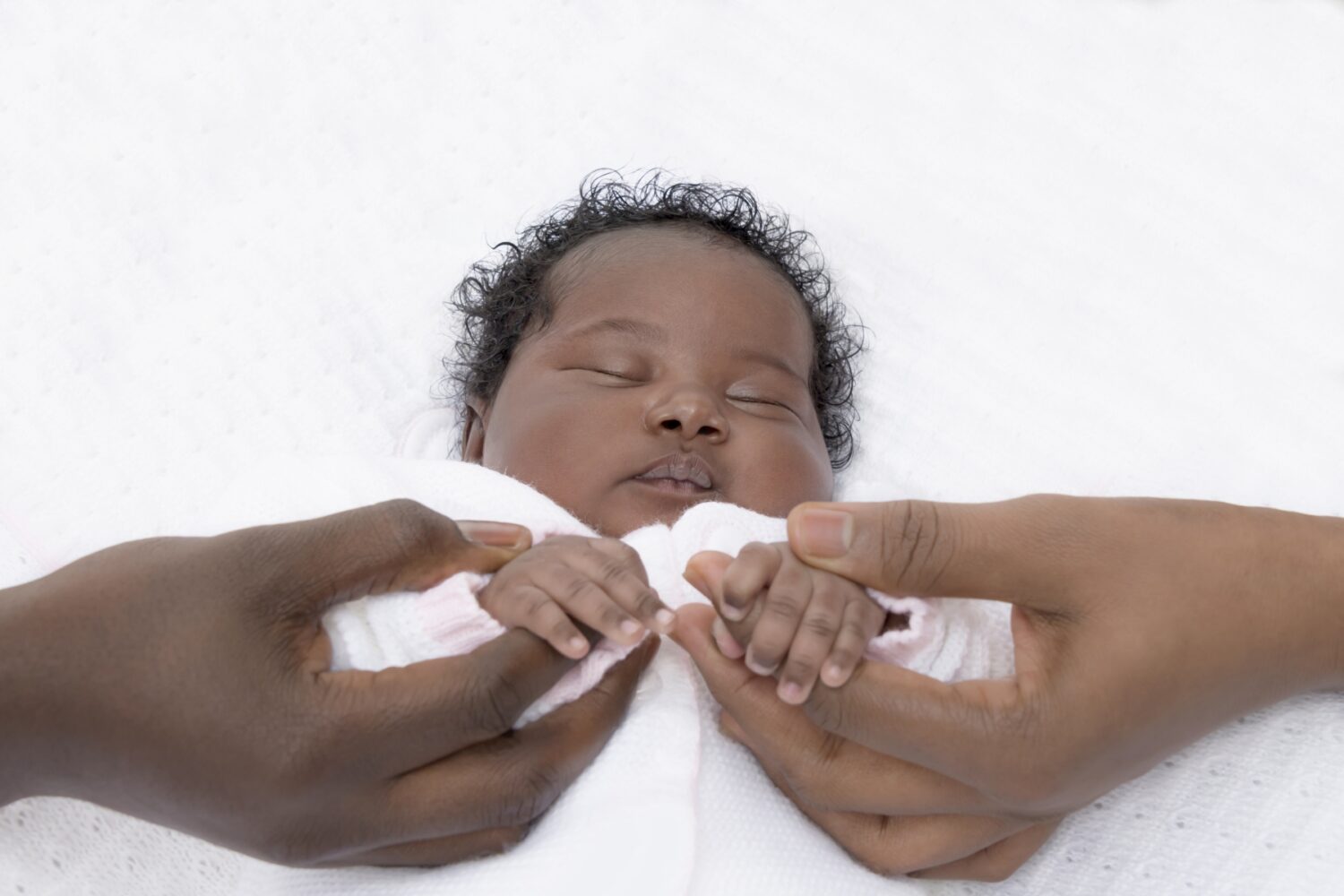 Two adult hands hold the hands of a tiny baby lying sleeping